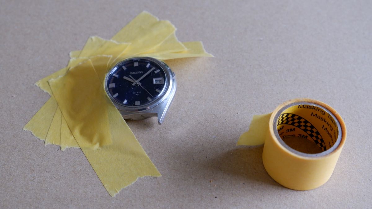 A watch with strap removed and half-covered in masking tape, exposing just the acrylic crystal.